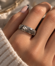 A close-up of a hand wearing a textured silver band ring with engraved details, styled with a cream knit sweater and warm natural sunlight.