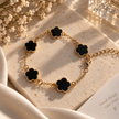 A gold chain bracelet with five black clover-shaped charms, styled on a textured beige stone with white flowers and cream satin fabric.