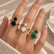 A close-up of a hand wearing four gold-tone clover-shaped rings with different enamel fills: two black, one white mother-of-pearl, and one green striped. The hand has a French manicure and is wearing a cream knit sweater.