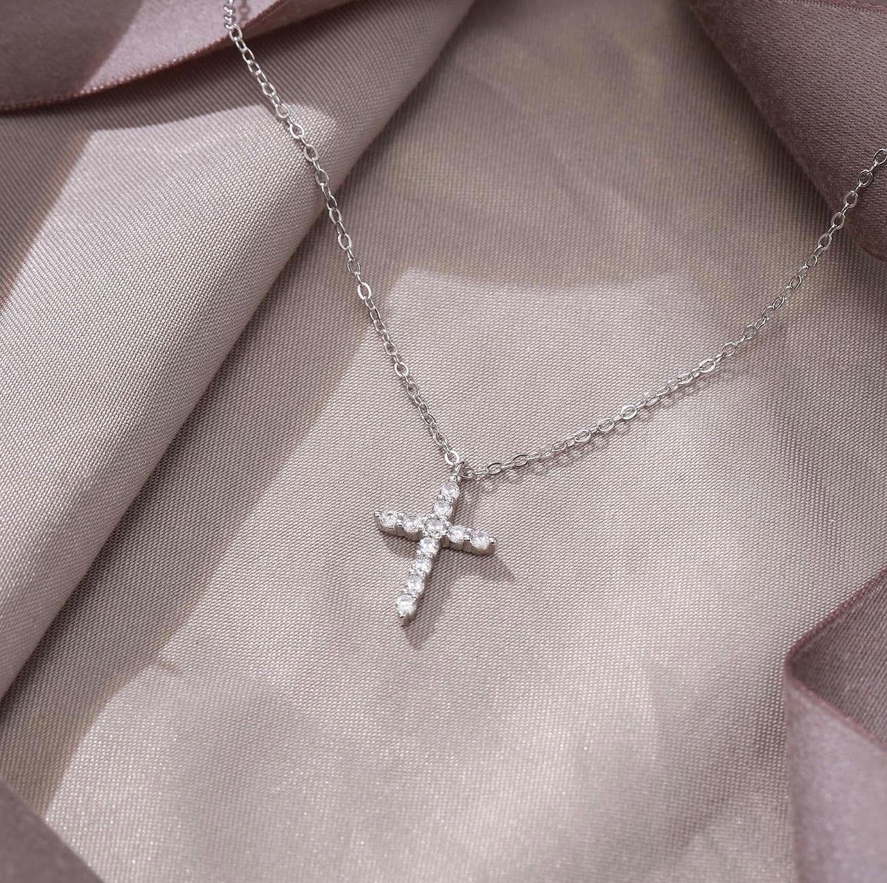 A silver cross pendant necklace adorned with small clear stones, resting on draped champagne-colored satin fabric.