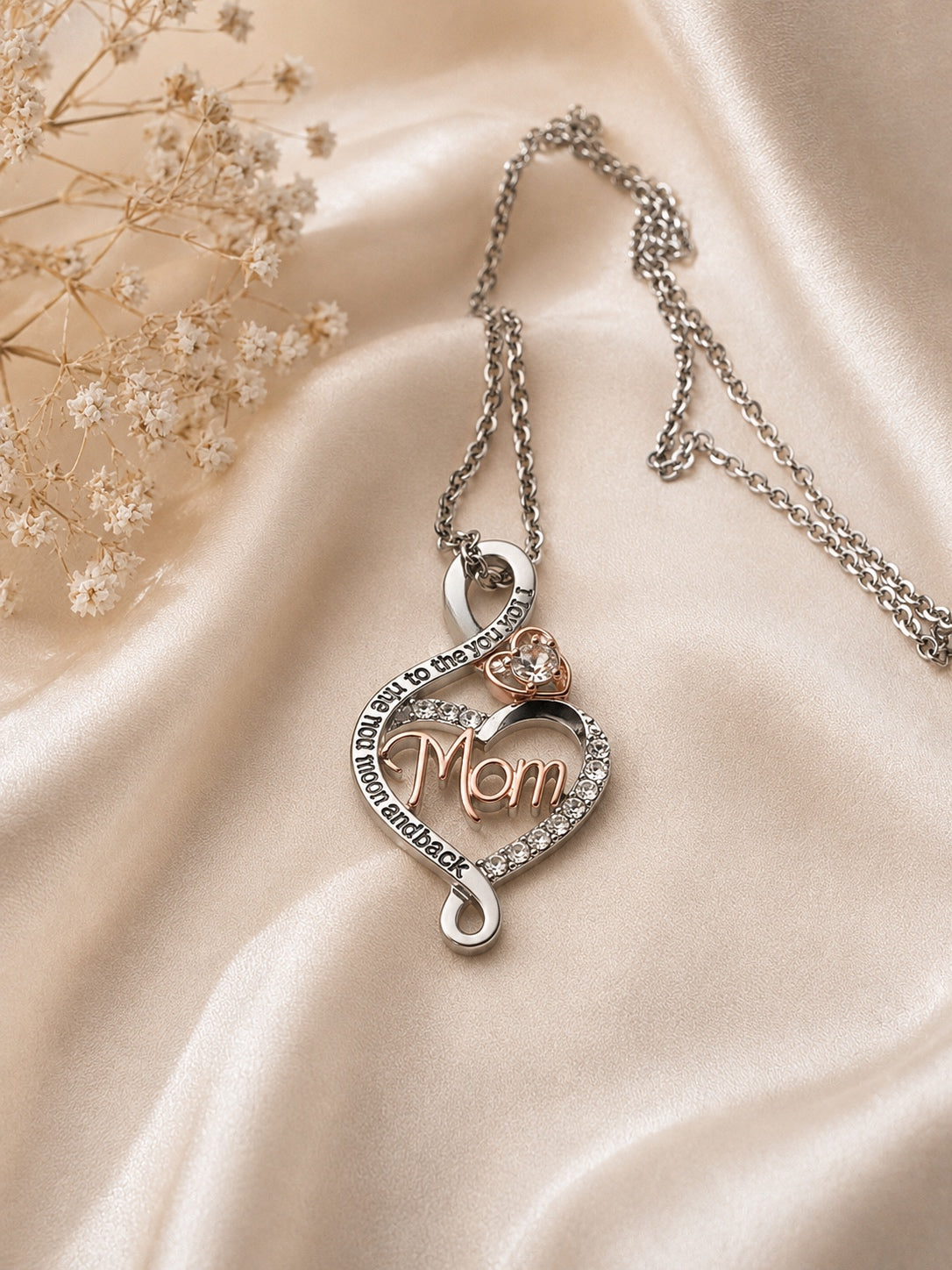 A silver-toned necklace with a heart-shaped pendant featuring the word 'Mom' in rose gold, surrounded by small clear stones and a small heart-shaped stone accent, displayed on draped champagne satin fabric with dried flowers.
