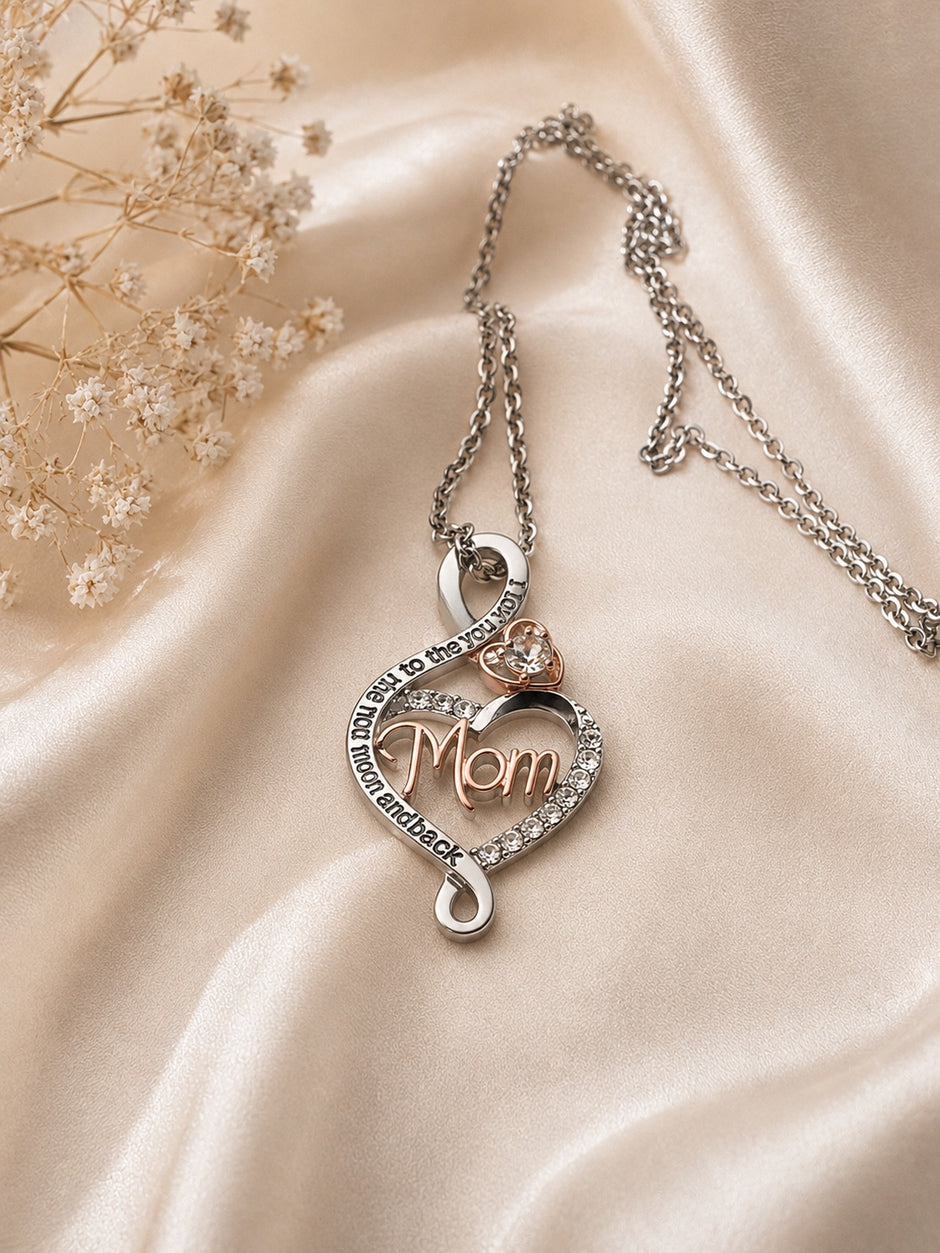 A silver-toned necklace with a heart-shaped pendant featuring the word 'Mom' in rose gold, surrounded by small clear stones and a small heart-shaped stone accent, displayed on draped champagne satin fabric with dried flowers.