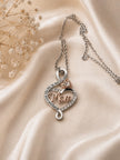 A silver-toned necklace with a heart-shaped pendant featuring the word 'Mom' in rose gold, surrounded by small clear stones and a small heart-shaped stone accent, displayed on draped champagne satin fabric with dried flowers.