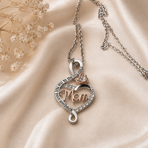 A silver-toned necklace with a heart-shaped pendant featuring the word 'Mom' in rose gold, surrounded by small clear stones and a small heart-shaped stone accent, displayed on draped champagne satin fabric with dried flowers.