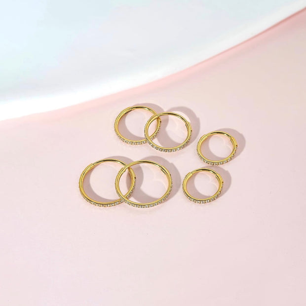 Five gold rings with small clear stones set along the outer edges, arranged on a soft pink background with a white curved shape in the upper left.