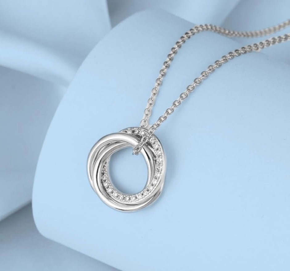A silver-toned necklace with a double interlocking ring pendant, accented with small clear stones along one of the rings, displayed on a light blue jewelry bust.