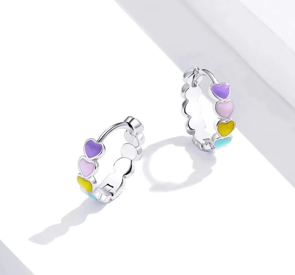 A pair of silver huggie hoop earrings decorated with a row of pastel-colored heart charms in purple, pink, yellow, and blue, displayed on a clean white surface.