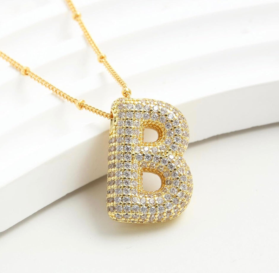 Gold letter B pendant necklace paved with clear rhinestones, resting on a white background.