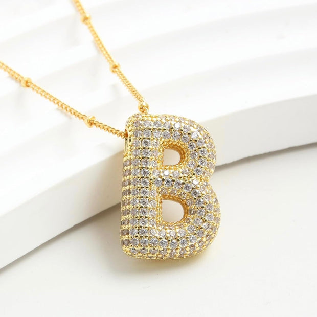 Gold letter B pendant necklace paved with clear rhinestones, resting on a white background.
