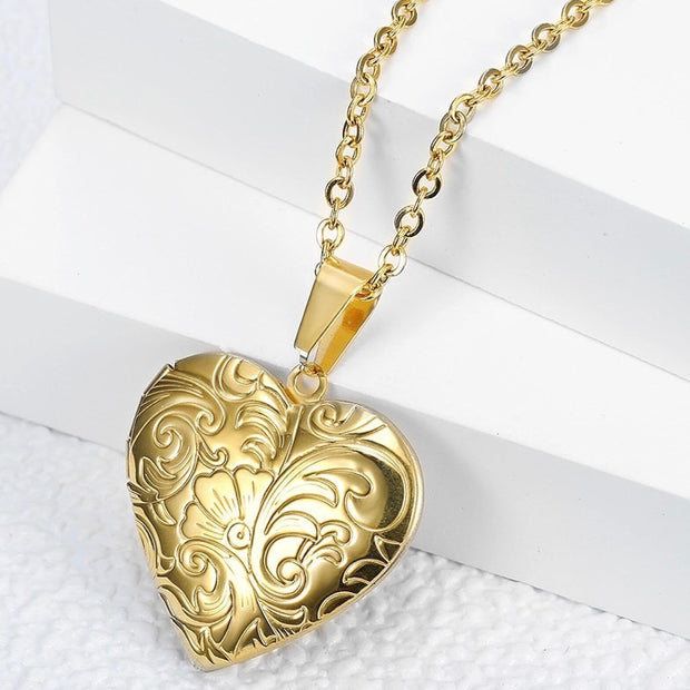 A gold heart-shaped locket pendant necklace with an ornate embossed floral pattern, hanging from a gold chain against a white geometric background.