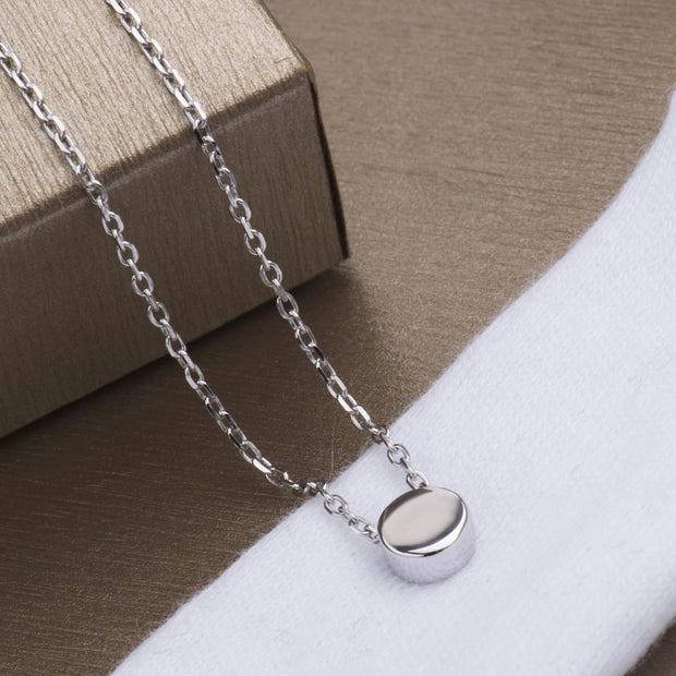 A silver chain necklace with a small, polished oval pendant resting on a white textured surface next to a brown box.