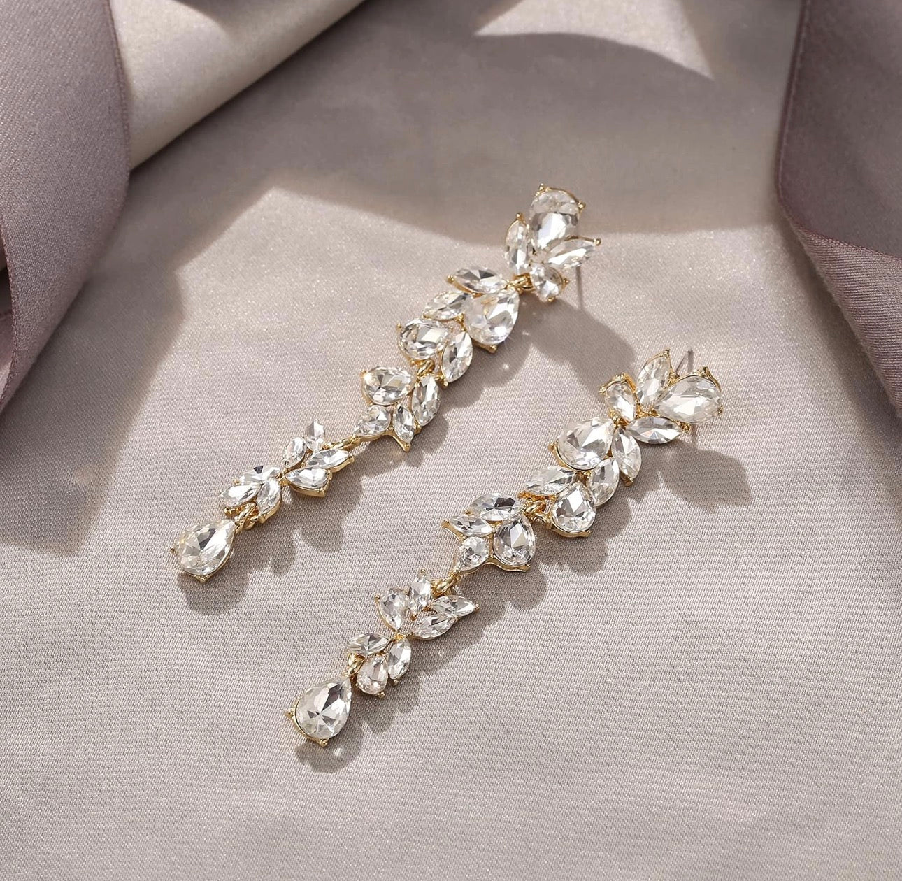 A pair of gold-toned drop earrings adorned with clear faceted crystals arranged in a leafy vine pattern, displayed on a soft beige fabric background.