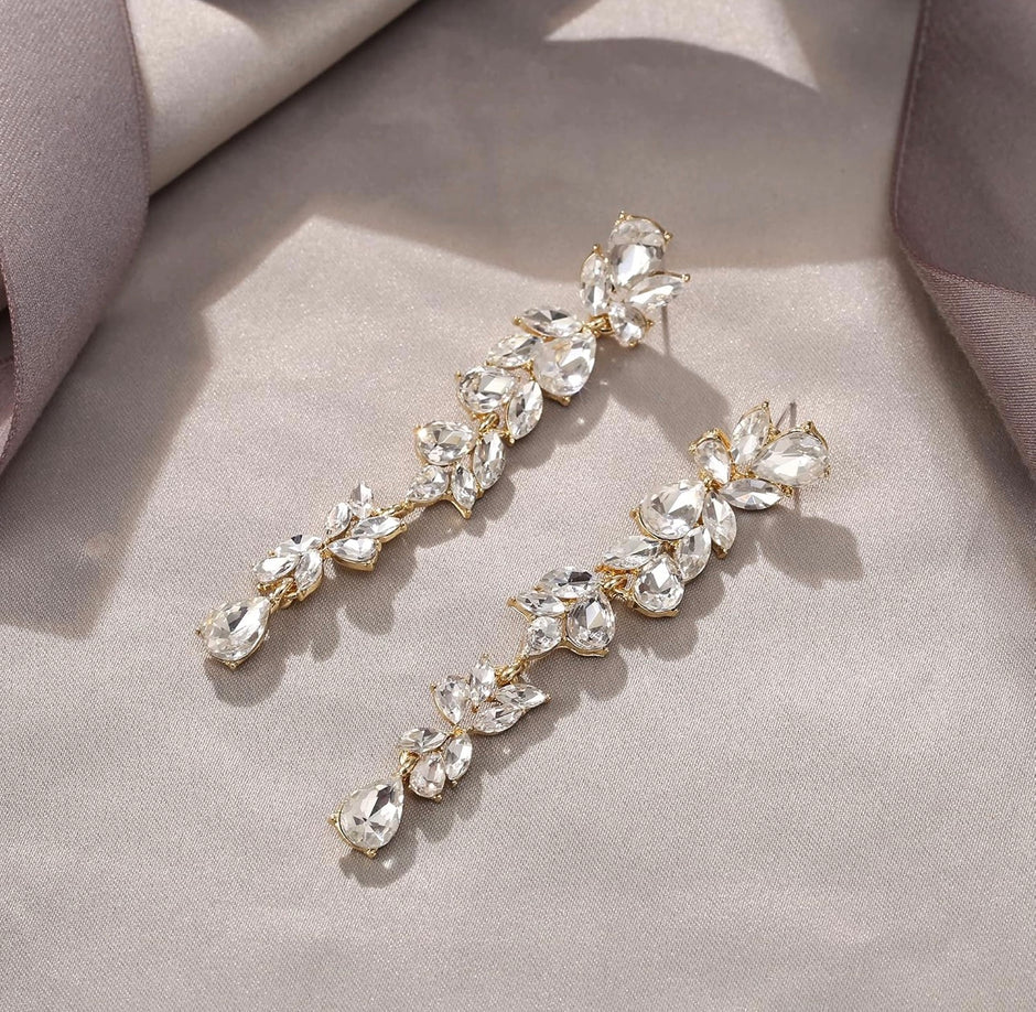 A pair of gold-toned drop earrings adorned with clear faceted crystals arranged in a leafy vine pattern, displayed on a soft beige fabric background.