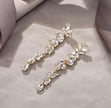 A pair of gold-toned drop earrings adorned with clear faceted crystals arranged in a leafy vine pattern, displayed on a soft beige fabric background.