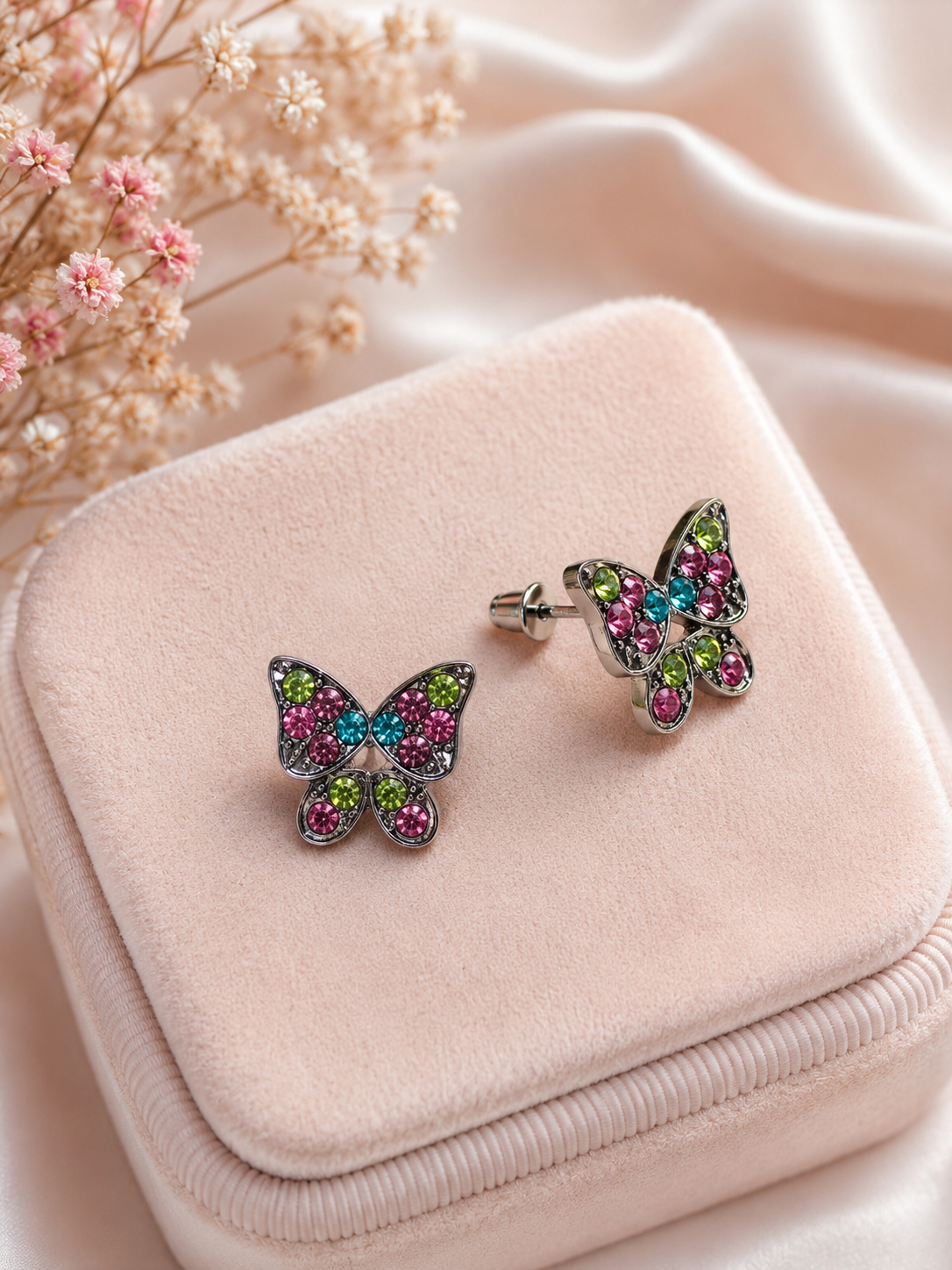 A pair of butterfly-shaped stud earrings with silver-toned metal and pink, green, and blue rhinestones, displayed on a light pink velvet jewelry box with dried flowers in the background.