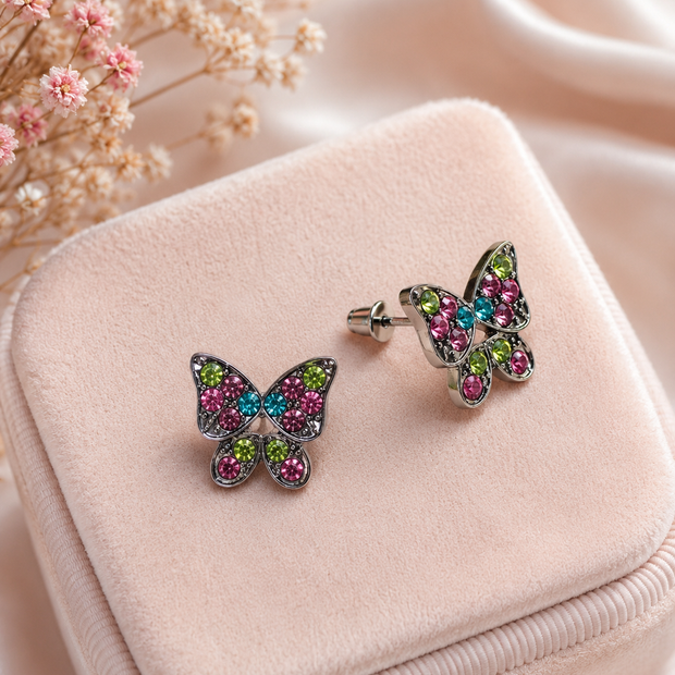A pair of butterfly-shaped stud earrings with silver-toned metal and pink, green, and blue rhinestones, displayed on a light pink velvet jewelry box with dried flowers in the background.