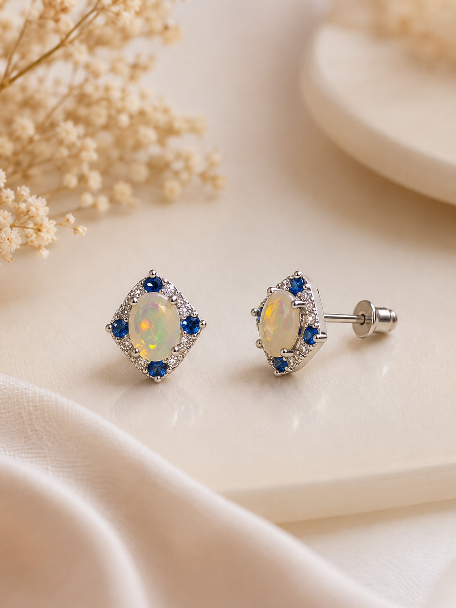 A pair of diamond-shaped stud earrings with iridescent opal centers and blue accent stones in a silver setting, displayed on a cream-colored surface with dried flowers.