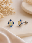 A pair of diamond-shaped stud earrings with iridescent opal centers and blue accent stones in a silver setting, displayed on a cream-colored surface with dried flowers.