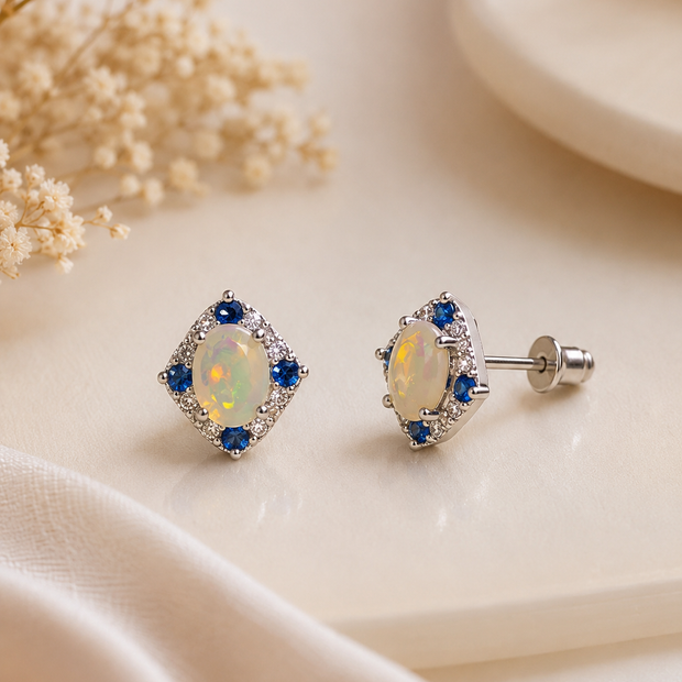 A pair of diamond-shaped stud earrings with iridescent opal centers and blue accent stones in a silver setting, displayed on a cream-colored surface with dried flowers.