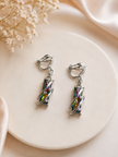 A pair of silver-toned clip-on earrings with iridescent multicolor geometric charms, displayed on a round cream ceramic plate with soft satin and dried flowers in the background.