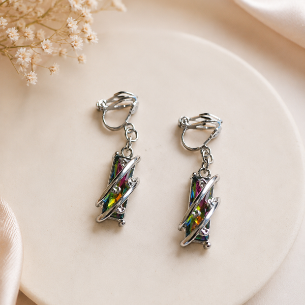 A pair of silver-toned clip-on earrings with iridescent multicolor geometric charms, displayed on a round cream ceramic plate with soft satin and dried flowers in the background.