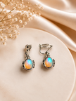 A pair of silver-toned drop earrings with oval iridescent opal-like stones, displayed on a round cream surface with soft satin fabric and dried flowers in the background.