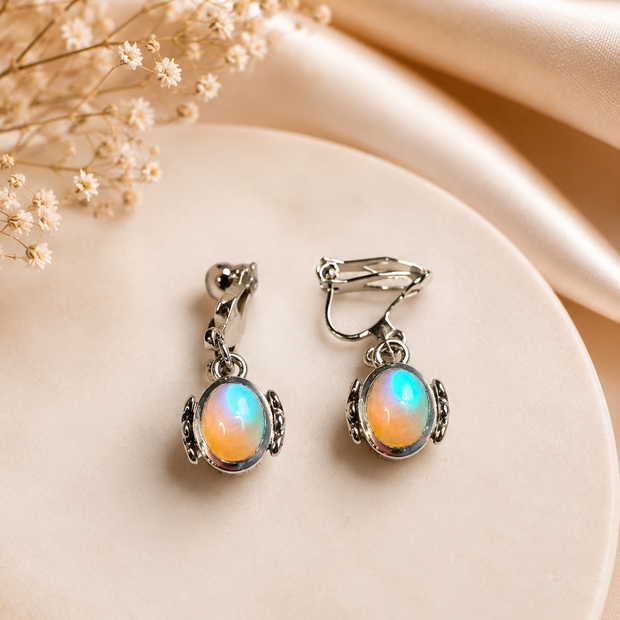 A pair of silver-toned drop earrings with oval iridescent opal-like stones, displayed on a round cream surface with soft satin fabric and dried flowers in the background.