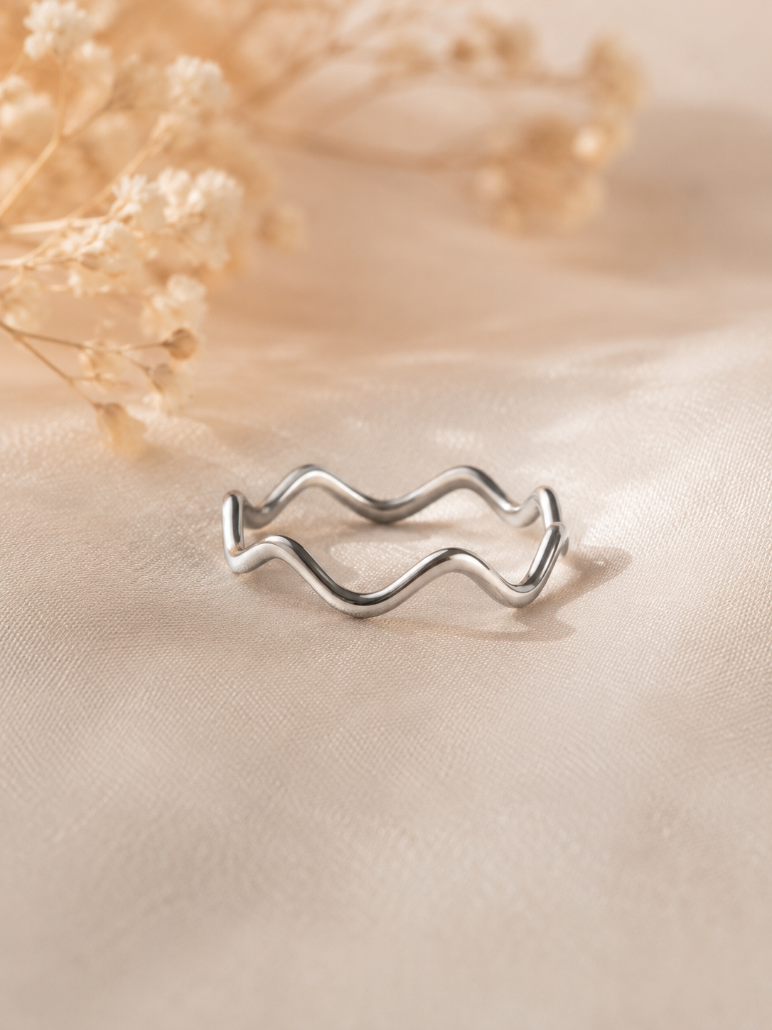 A silver wavy open band ring resting on a beige satin fabric surface with dried flowers in the background.