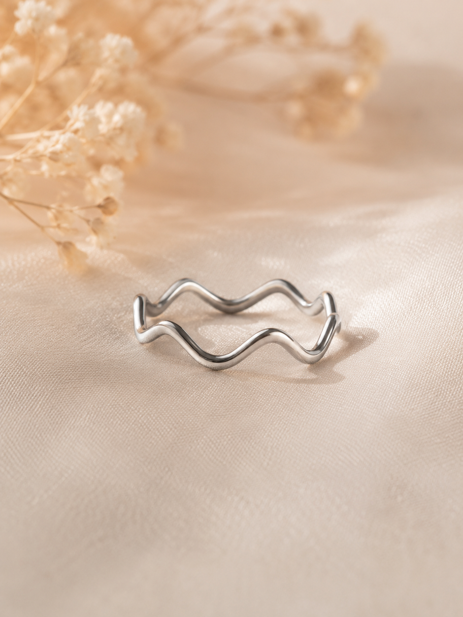 A silver wavy open band ring resting on a beige satin fabric surface with dried flowers in the background.