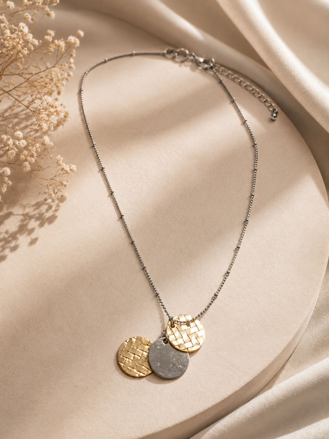 A delicate silver chain necklace with three overlapping round pendant discs in textured gold, smooth gold, and matte gray, displayed on a beige satin surface with dried flowers.