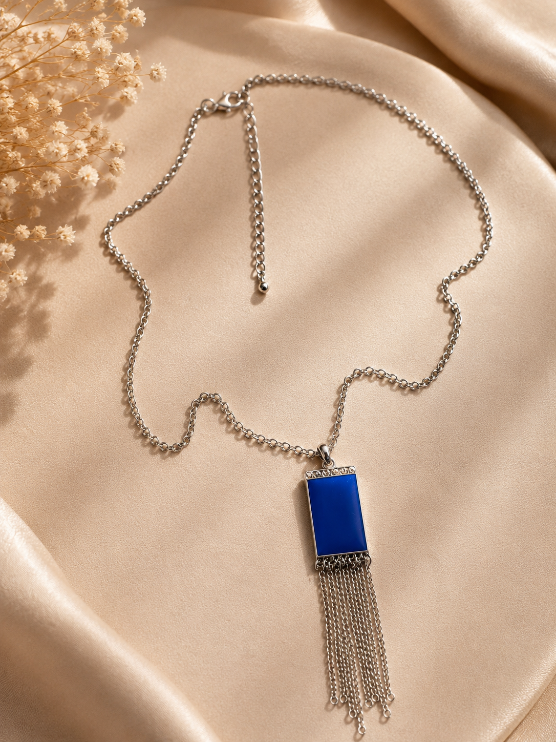 A silver-toned chain necklace with a rectangular royal blue pendant and dangling chain fringe, styled on draped beige satin fabric with dried flowers in the corner.