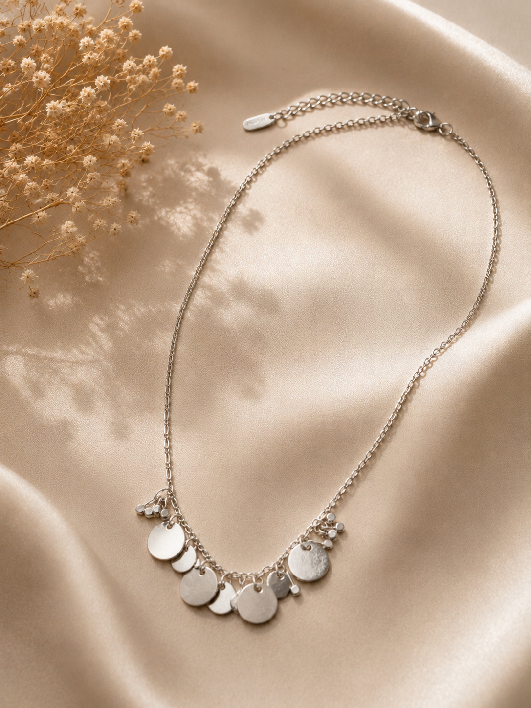 A silver-toned chain necklace with multiple flat round disc charms and small bead accents, arranged on draped satin fabric with dried flowers in the corner.