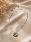 A silver chain necklace with a double interlocking ring pendant, decorated with small clear crystals, displayed on a beige satin fabric background with dried flowers in the corner.