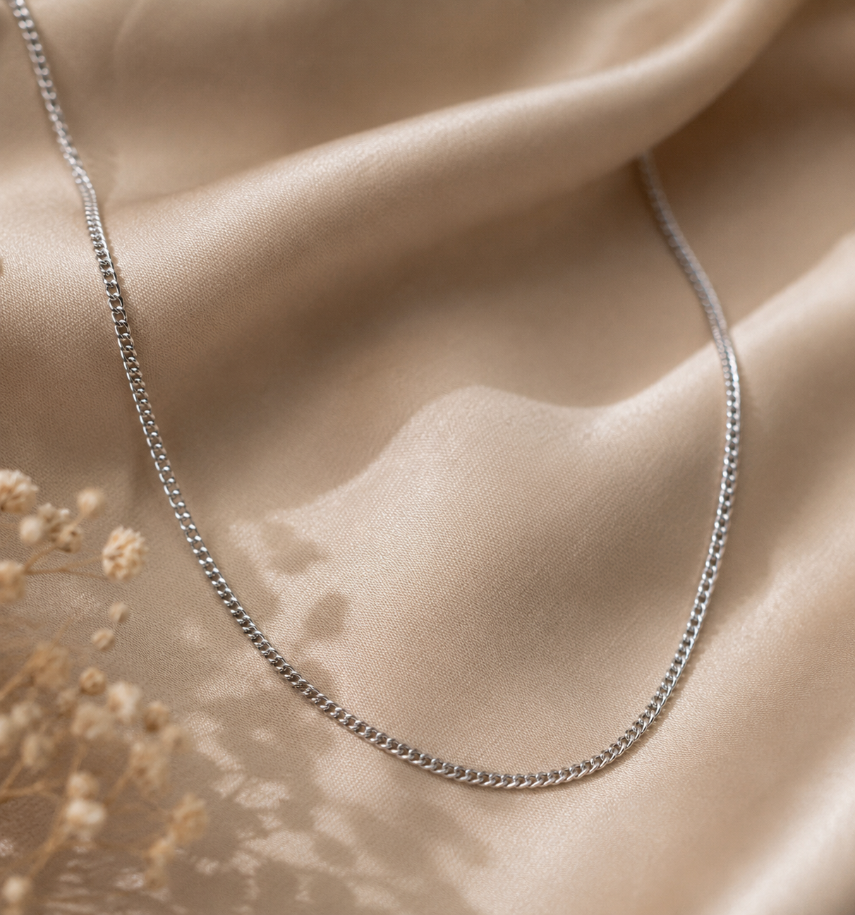 A silver chain necklace draped elegantly over draped champagne-colored satin fabric with dried flowers in the corner.