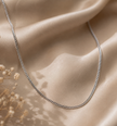 A silver chain necklace draped elegantly over draped champagne-colored satin fabric with dried flowers in the corner.