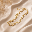 A gold paperclip chain necklace with a matching extender chain, styled on draped beige satin fabric with dried baby's breath flowers in the corner.