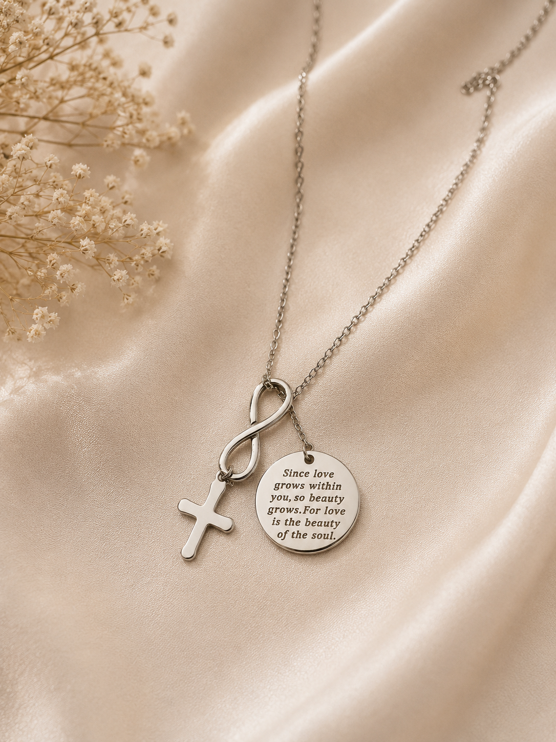 A silver-toned necklace with three pendants: a cross, an infinity symbol, and a round engraved disc, displayed on draped satin fabric with dried flowers.