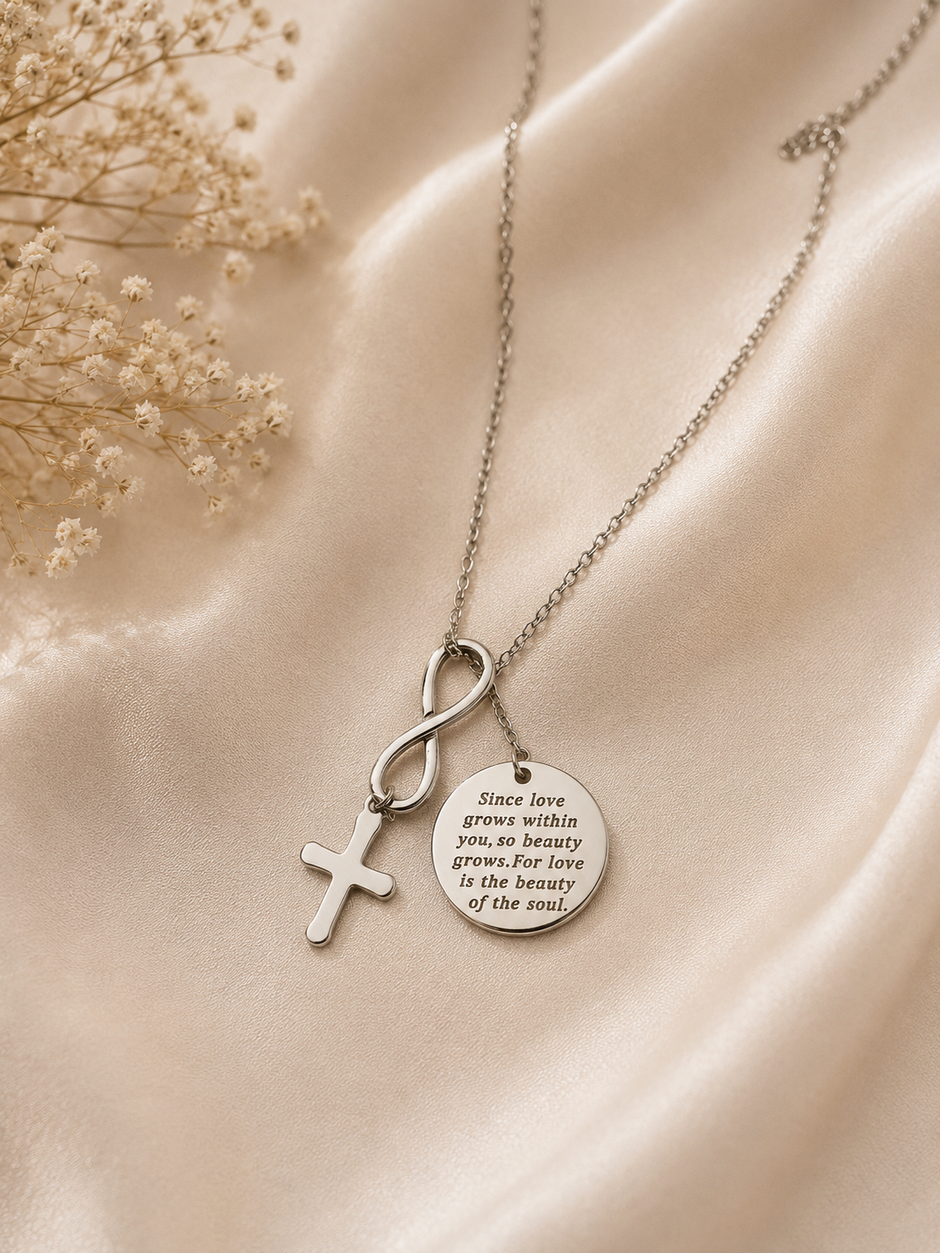 A silver-toned necklace with three pendants: a cross, an infinity symbol, and a round engraved disc, displayed on draped satin fabric with dried flowers.
