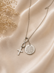A silver-toned necklace with three pendants: a cross, an infinity symbol, and a round engraved disc, displayed on draped satin fabric with dried flowers.