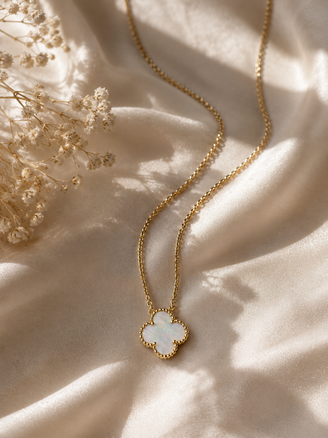 A gold chain necklace with a clover-shaped pendant featuring a white iridescent inlay, styled on draped cream satin fabric with dried flowers.