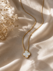 A gold chain necklace with a clover-shaped pendant featuring a white iridescent inlay, styled on draped cream satin fabric with dried flowers.