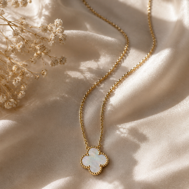 A gold chain necklace with a clover-shaped pendant featuring a white iridescent inlay, styled on draped cream satin fabric with dried flowers.