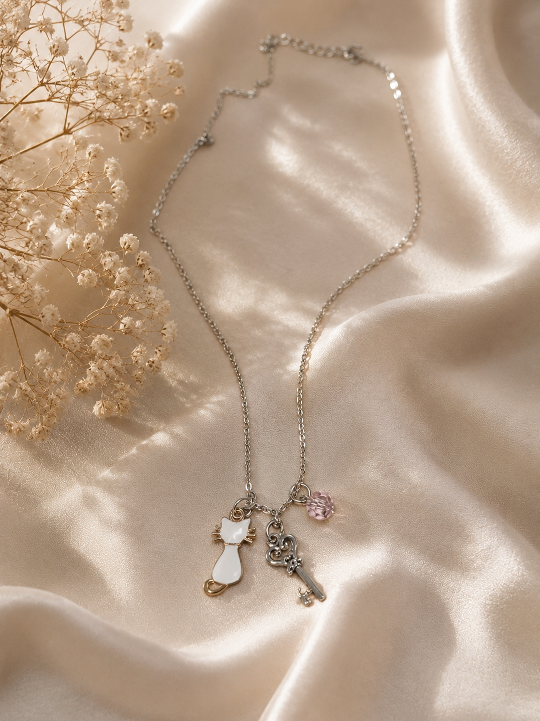 A delicate silver-toned key necklace with a white cat charm, a silver key charm, and a small pink gem accent, displayed on draped champagne satin fabric with dried baby's breath flowers.