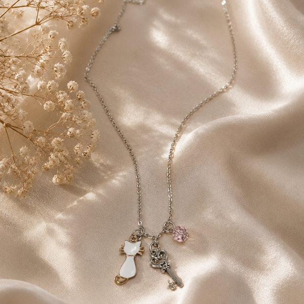 A delicate silver-toned key necklace with a white cat charm, a silver key charm, and a small pink gem accent, displayed on draped champagne satin fabric with dried baby's breath flowers.