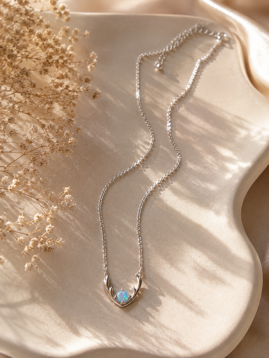 A delicate silver chain necklace with a crescent moon pendant holding a small light blue opal, displayed on a beige satin surface with dried flowers and soft shadows.