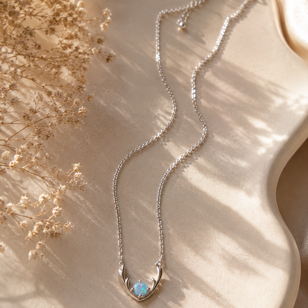 A delicate silver chain necklace with a crescent moon pendant holding a small light blue opal, displayed on a beige satin surface with dried flowers and soft shadows.