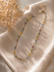 A delicate gold chain necklace adorned with small, colorful gemstone beads in yellow, blue, green, and purple, styled on draped champagne satin fabric with dried baby's breath flowers.