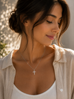A woman wearing a delicate silver cross pendant necklace, styled with a white ribbed tank top and a light beige linen button-up shirt. The photo is taken in warm, natural sunlight with soft shadows on a light-colored wall.