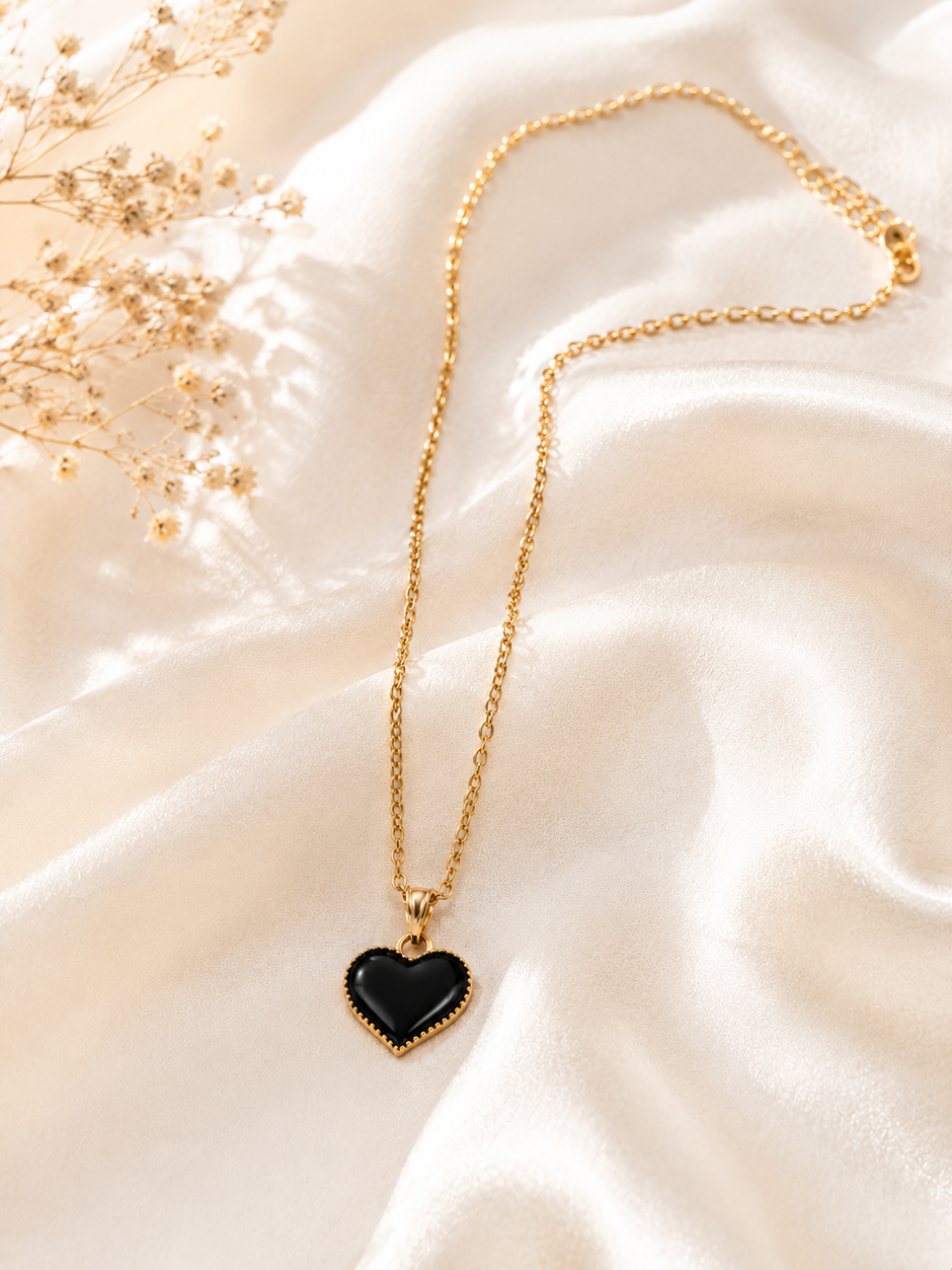A gold chain necklace with a heart-shaped pendant featuring a glossy black center and a gold border, displayed on draped cream satin fabric with dried floral accents.