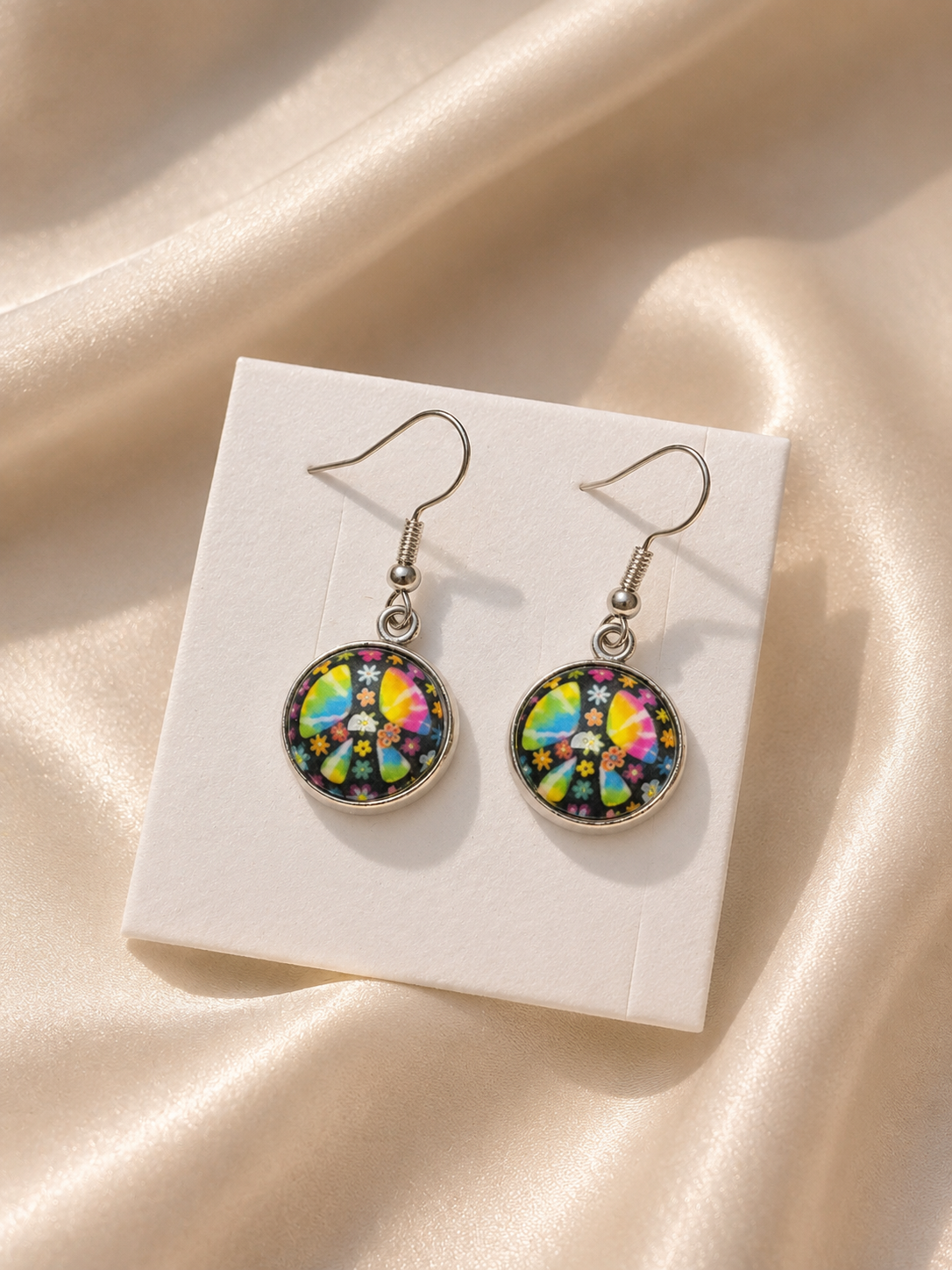 A pair of dangling earrings with colorful circular mandala designs in silver hooks, displayed on a white card over a beige satin fabric background.